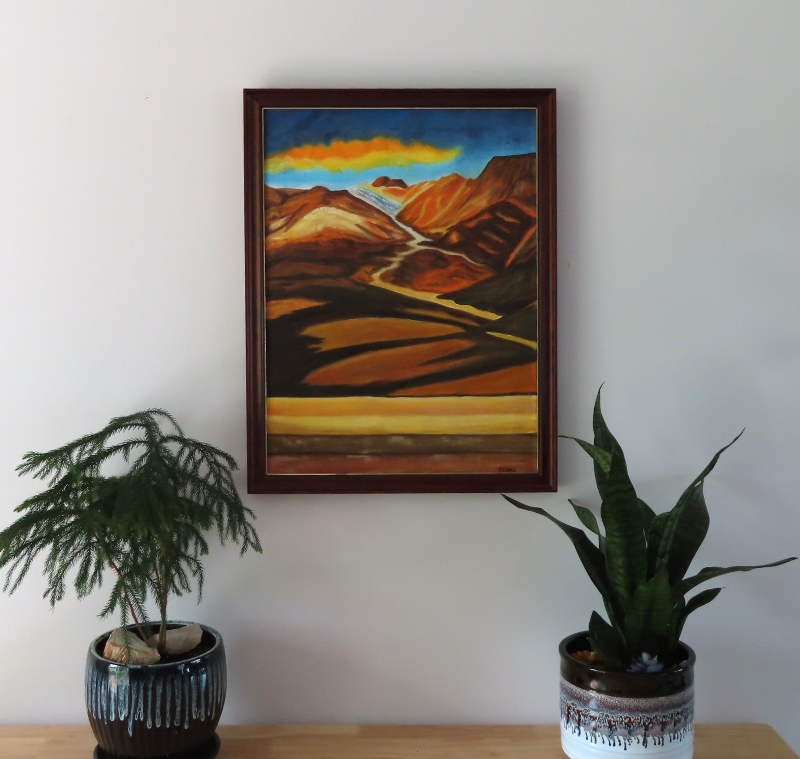 original oil painting of colorful mountains and plains with colorful clouds in sky framed in complementary wood frame, shown over table