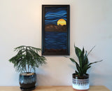 12 inches wide by 24 inches tall original oil painting, in an abstracted style, of a bright colored planet rising over the horizon in another world cool blue sky and water framed in black shown over table with potted plants