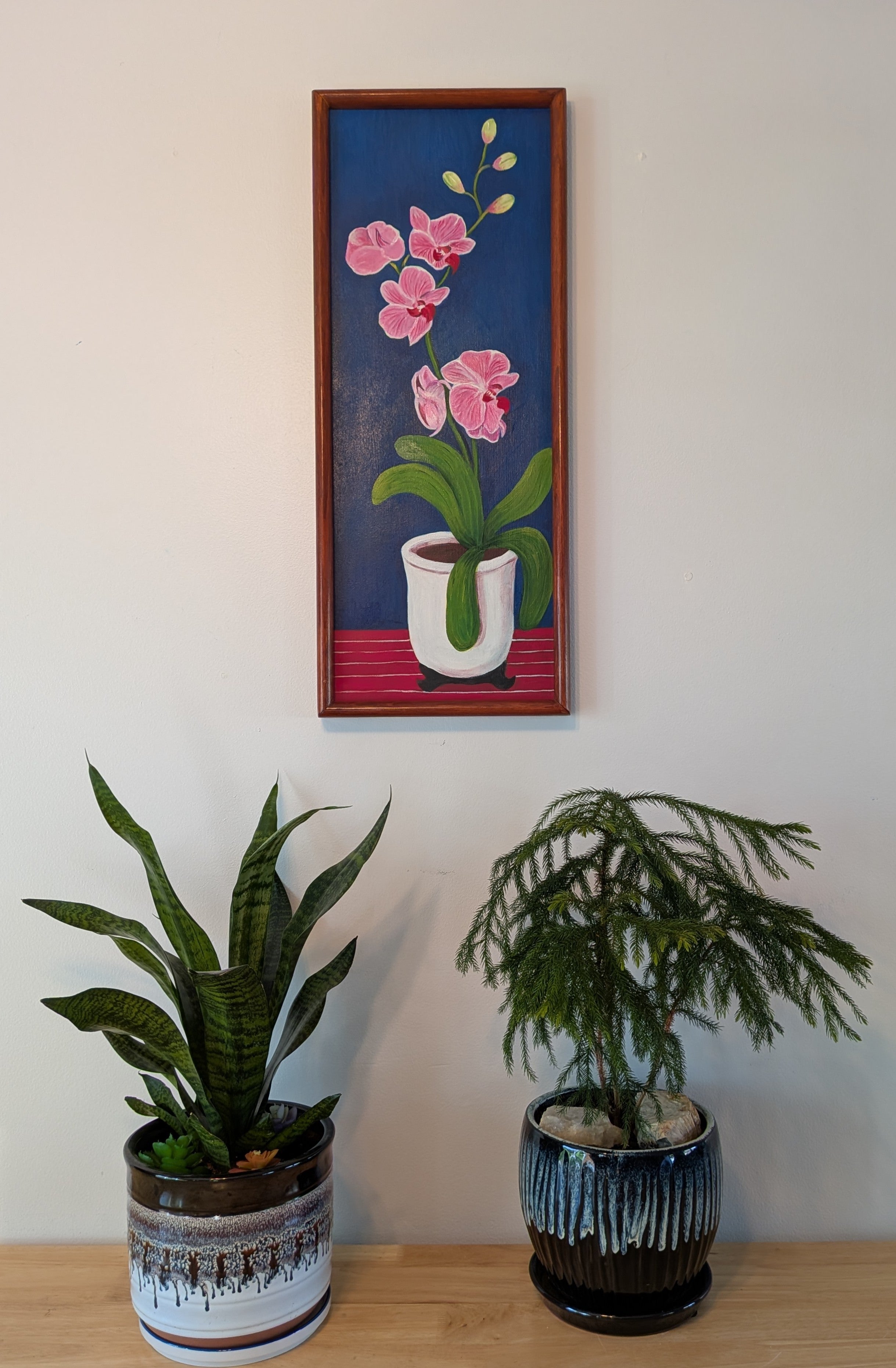 original oil painting on board of pink orchids against a royal blue background, in a white pot, framed in a handmade red chestnut colored frame, displayed over a table