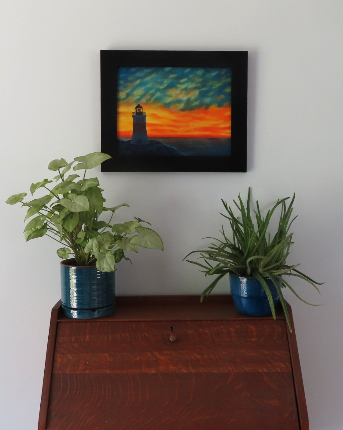 original oil painting of Erie Pierhead light house silhouetted by beautiful sunset and clouds, painted on 11 x 14 canvas panel, framed in modern black frame, displayed over secretary desk