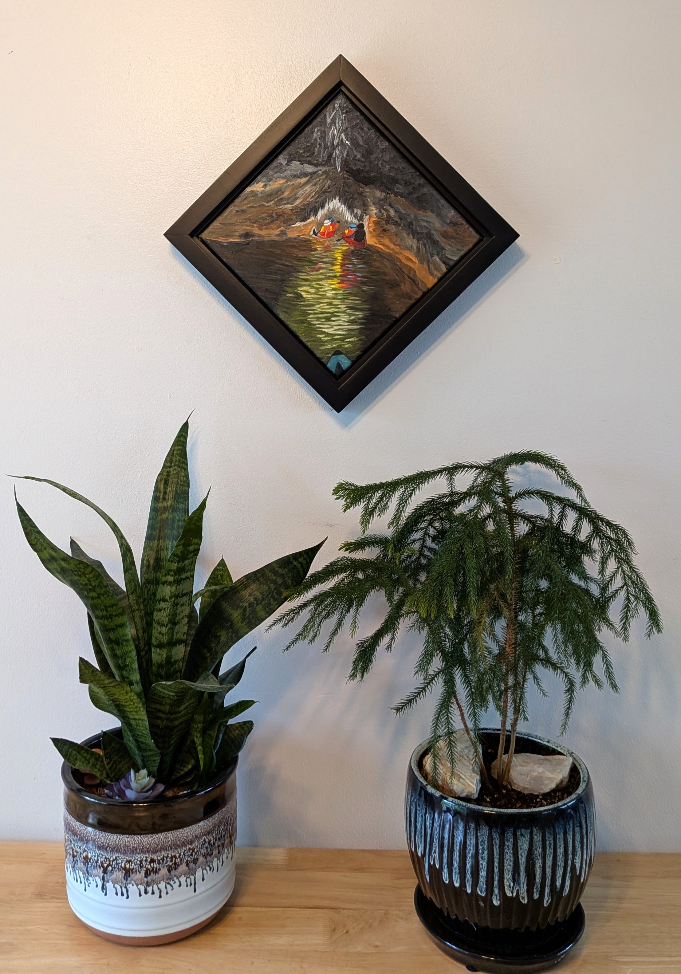 original oil painting of cavers in canoes entering a cave system with river, dark water and walls, accented by light reflections and cave formations, framed in black, diamond presentation, displayed over table