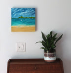 12x12" original oil painting on deep cradle canvas of turquoise ocean, sandy beach with sailboats and mountains in the distance, shown over secretary desk