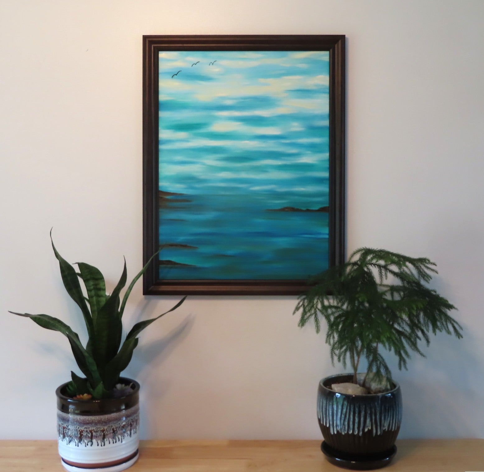 original oil painting of blue sky meeting the blue and teal ocean in the distance,framed in black, picured over table