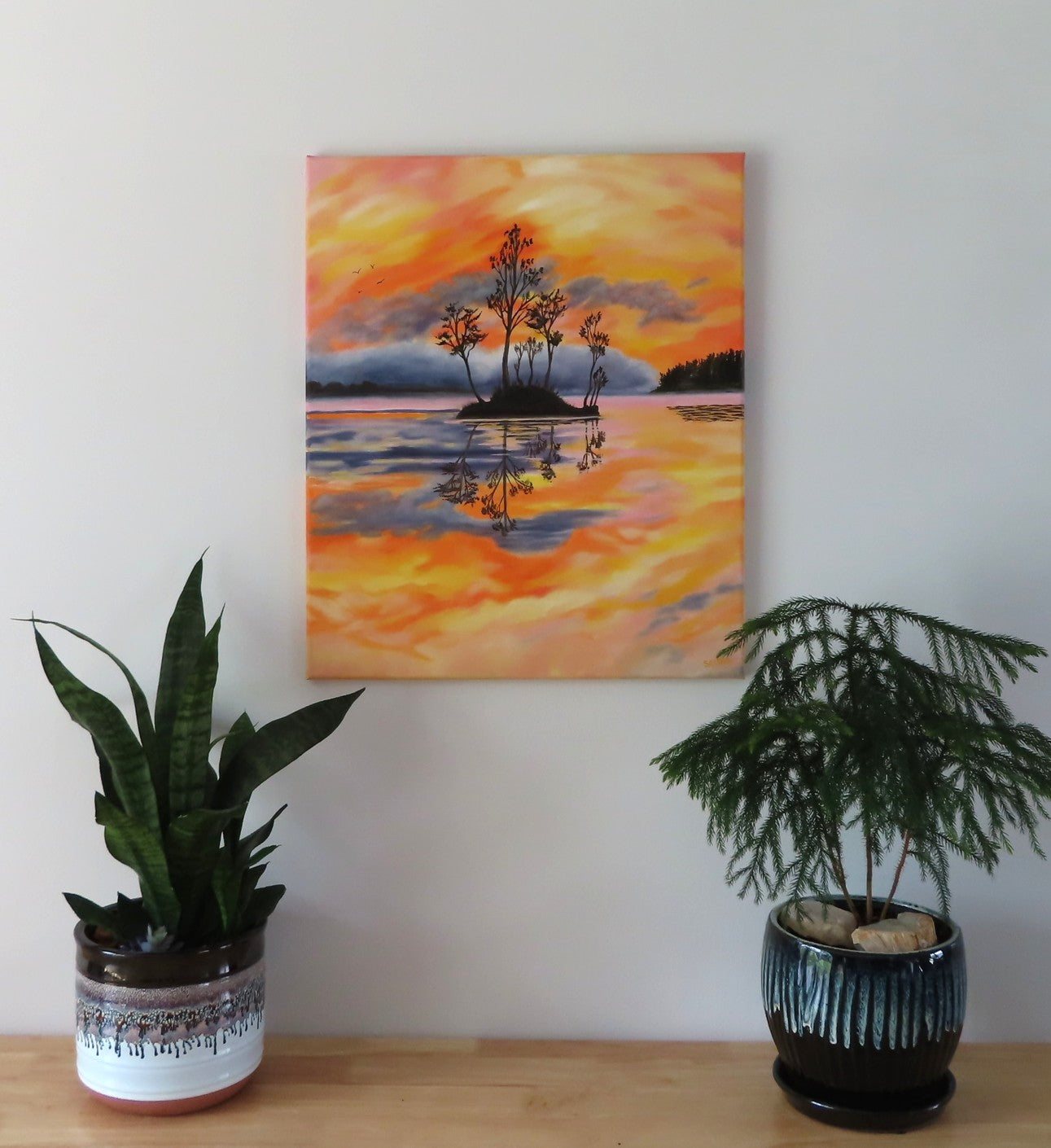 Original oil painting of a colorful pastel sky, the oranges, yellows and pinks contrast with the distant hills rising out of the mist and the dark silhouetted island and trees reflected in calm waters 24" tall by 20" wide unframed; shown over a table with plants