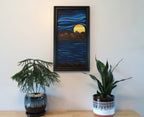 12x 24 vertical, original oil painting, in an abstracted style, of a bright colored planet rising over the horizon in another world cool blue sky and water framed in black shown over table with potted plants