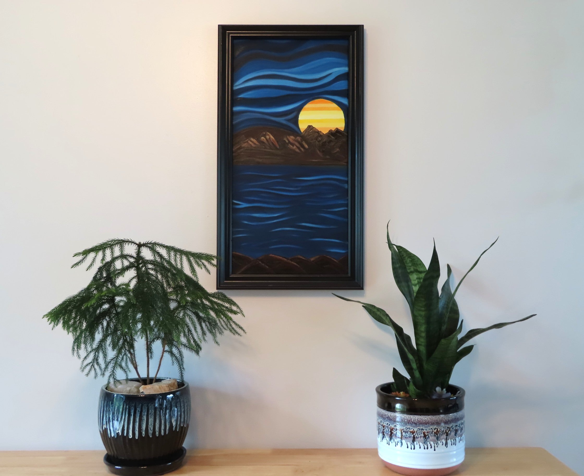 12x 24 vertical, original oil painting, in an abstracted style, of a bright colored planet rising over the horizon in another world cool blue sky and water framed in black shown over table with potted plants