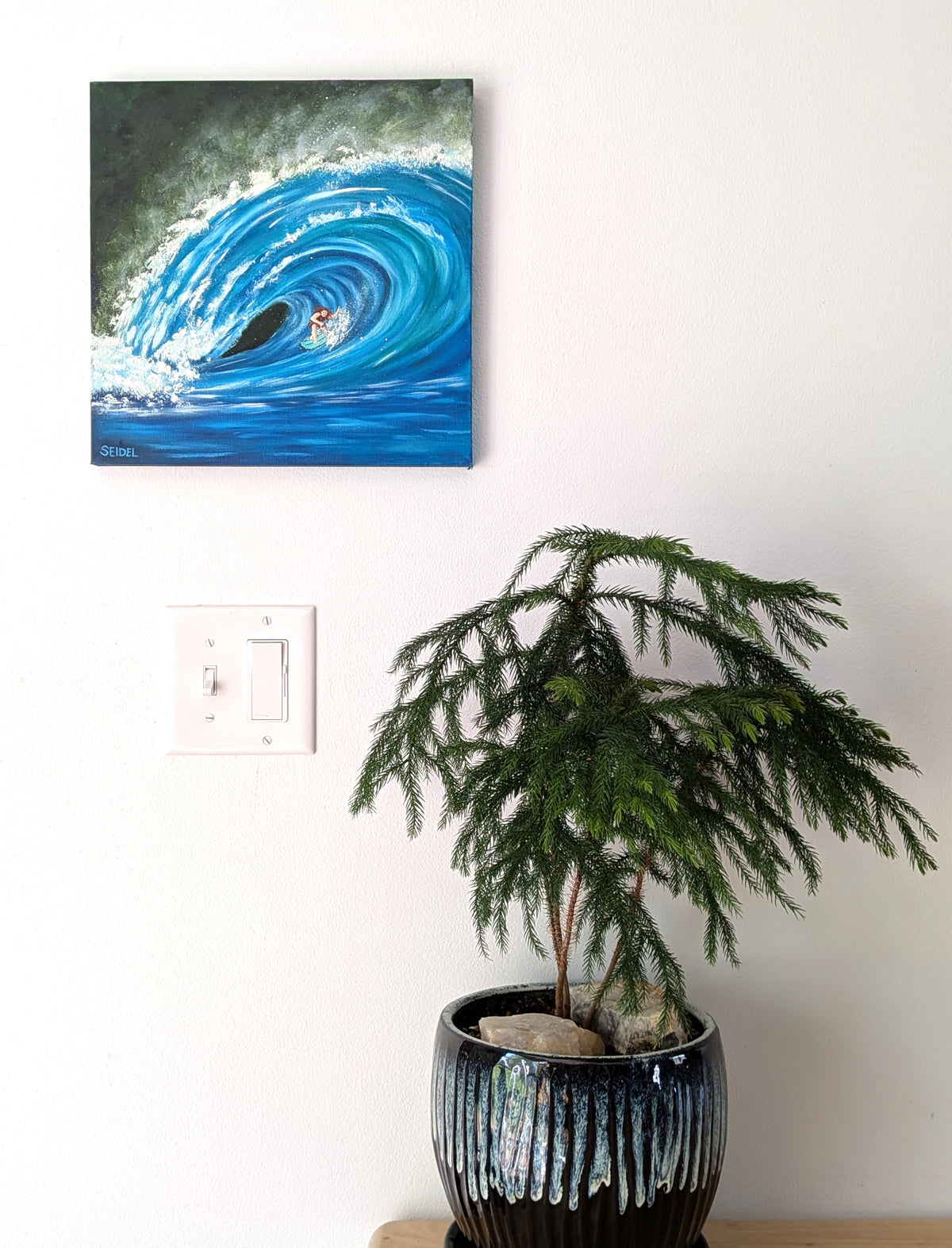 12x12" original oil painting of a surfer riding a curling blue ocean wave, displayed on a white wall above a potted green plant.